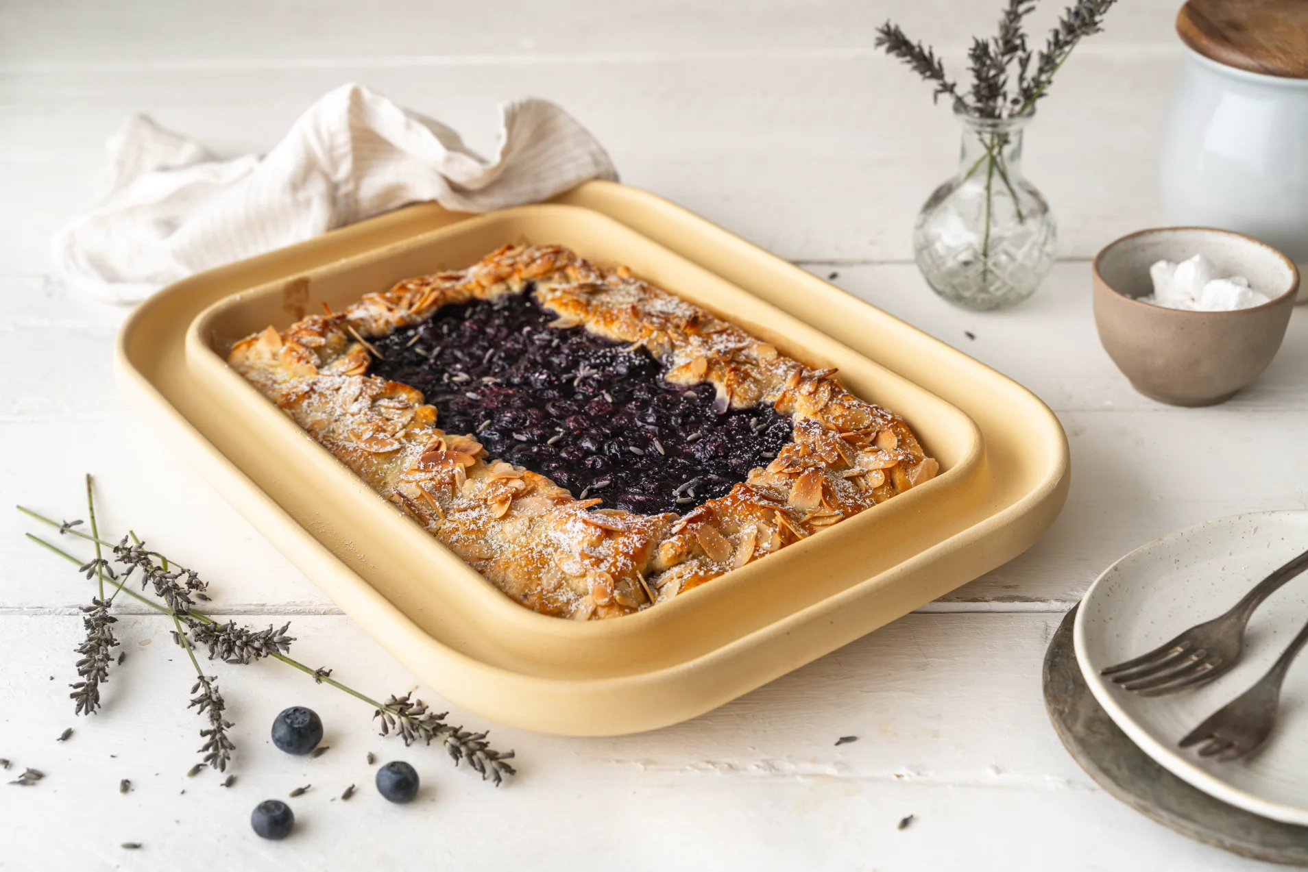 Ein köstlicher Blaubeerkuchen in einer beige-farbenen Backform mit goldener Kruste, verziert mit Mandelscheiben und einem Hauch von Puderzucker.